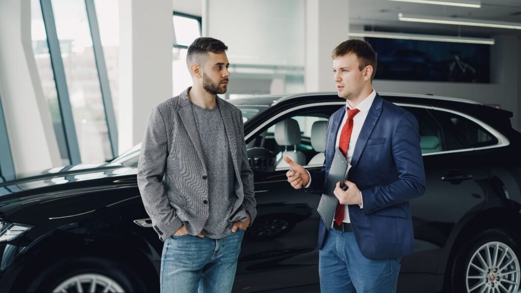 Two men discussing business in a car dealership, standing near a vehicle.